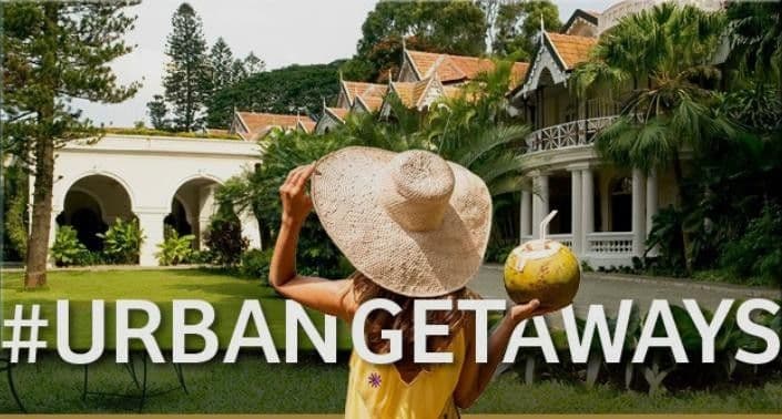 A woman in a sun hat holding a coconut in front of a luxury resort with the text “Urban Getaways".
