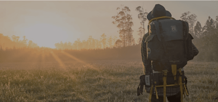 A person with a backpack walking through a grassy field at sunrise.